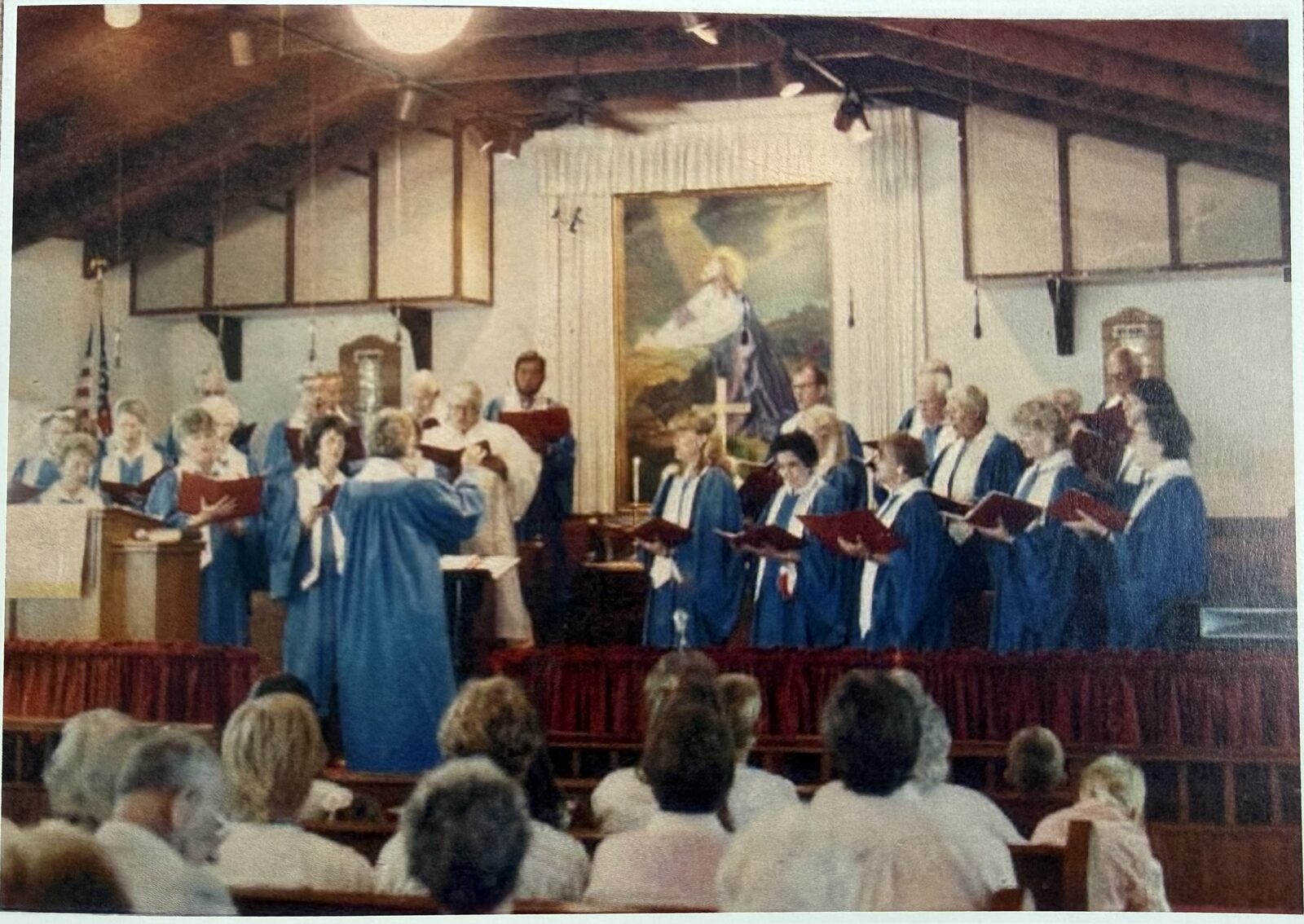 An older photo of the Big Pine choir in blue robes singing in the sanctuary, with Captain James Wilson's painting of Christ in Gethsemane behind them