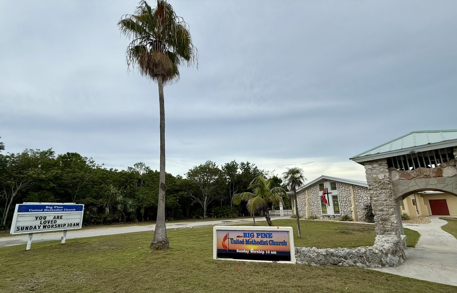 The front of Big Pine United Methodist Church from Key Deer Boulevard, with the church signs reading Sunday Worship 10 AM, the driveway, the sanctuary and the coral-rock bell tower