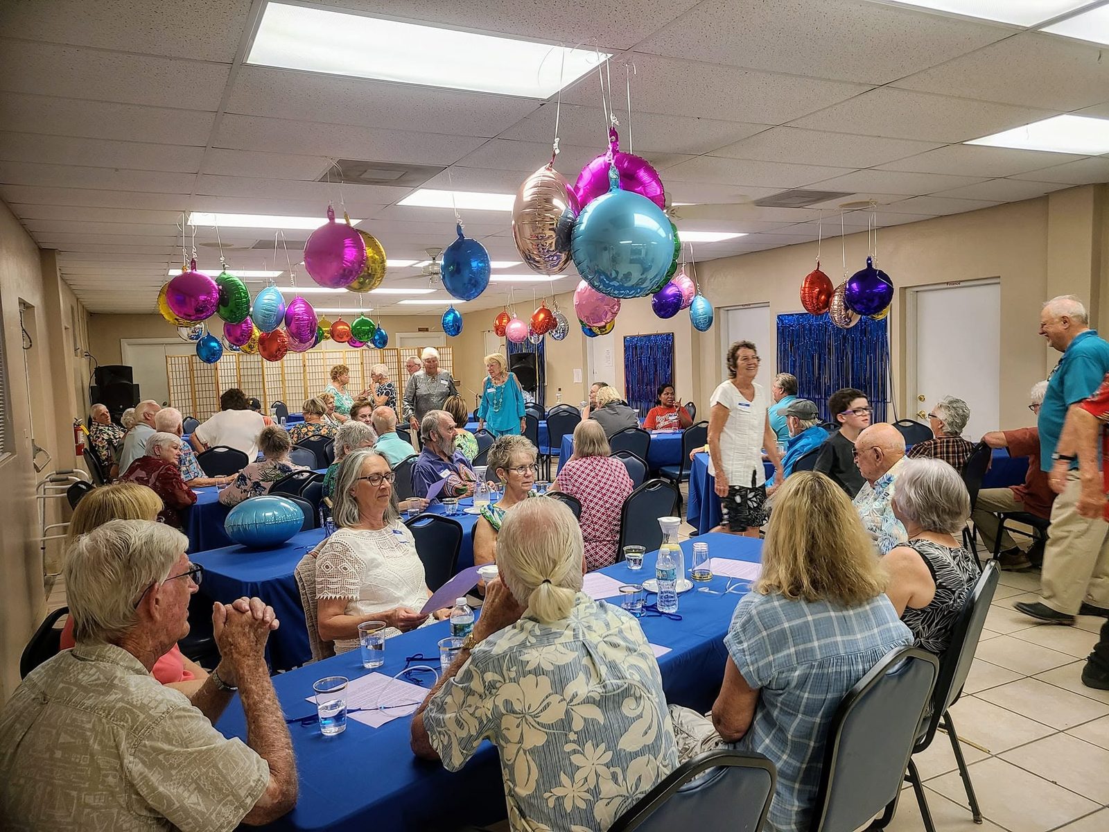Church members gathered around tables with blue tablecloths under colorful balloons in the fellowship hall