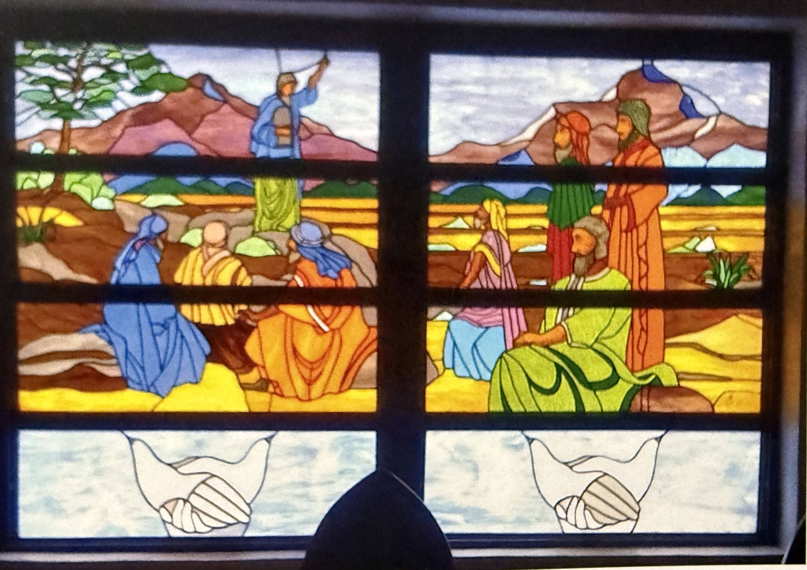 A colorful stained glass window designed by Bernal Johnson, showing a robed figure on a hillside raising tablets before a gathered crowd, with clasped hands in the lower panels